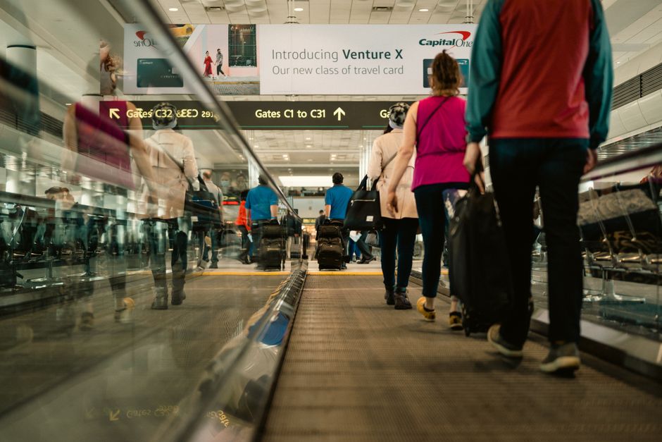 Passengers walking on a moving walkway in a busy airport terminal with signs for gates.