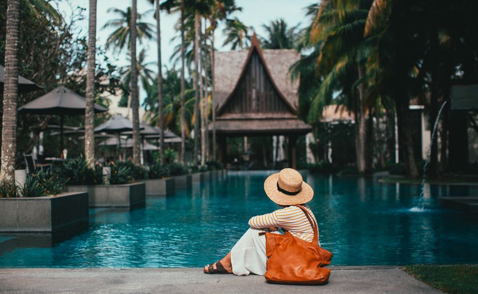 Side view anonymous female wearing summer clothes and hat resting on poolside in tropical resort amidst lush palms and bungalows