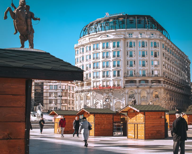 People walking near hotel and wooden booths in Skopje, showcasing urban architecture.
