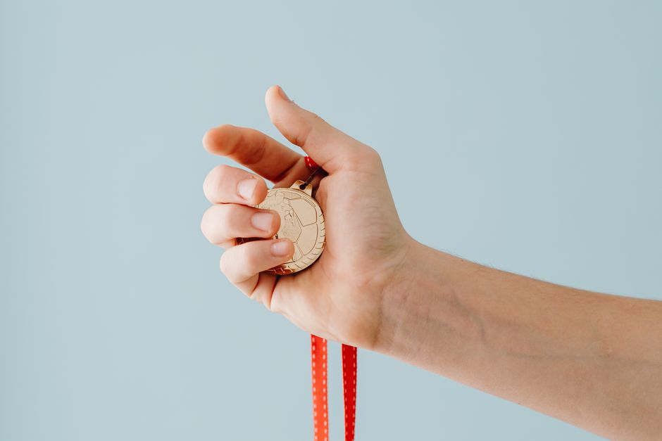 A close-up image of a hand holding a gold medal with a red ribbon against a blue background.