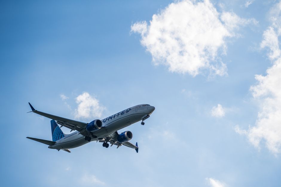 A United airplane flying high in the sky against a bright, cloudy backdrop.