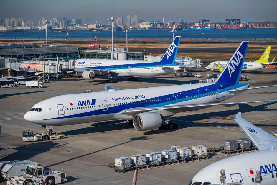 ANA Boeing 777 preparing for takeoff at Tokyo Haneda Airport with city skyline in background.