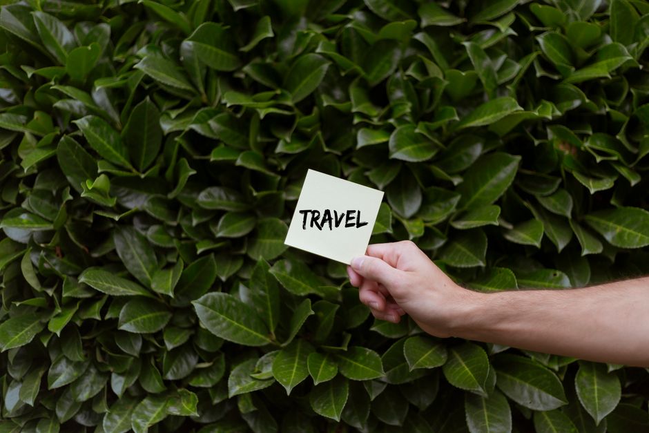A hand holds a note with the word 'travel' against a lush green bush background, embodying adventure.